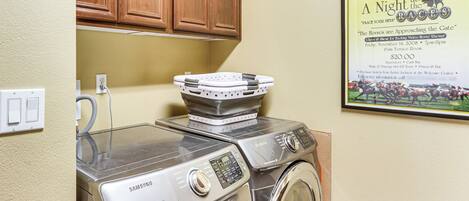 A laundry area with two Samsung washing machines, a wooden cabinet above, and a poster advertising a special event.