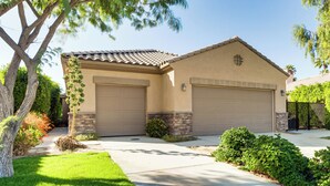 A two-story house with a beige exterior, two garage doors, and a well-maintained front yard with a tree and shrubs.