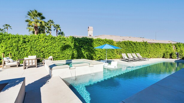 A swimming pool with lounge chairs and a blue umbrella, surrounded by a hedge and palm trees.
