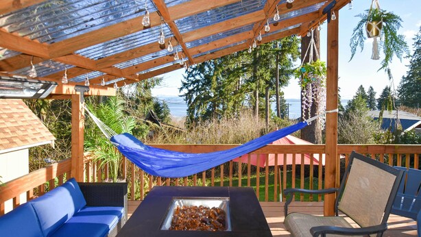 Eine Holzterrasse mit Glasdach, Sitzgelegenheiten im Freien, einer Feuerstelle und Blick auf den Ozean.