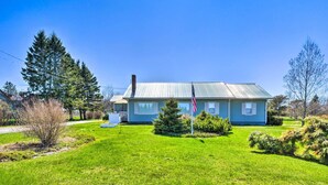 A two-story house with a blue exterior and a metal roof, surrounded by a well-maintained lawn and several trees. - Nautical Lubec Cottage w/ Fire Pit & Grill! (Lubec)