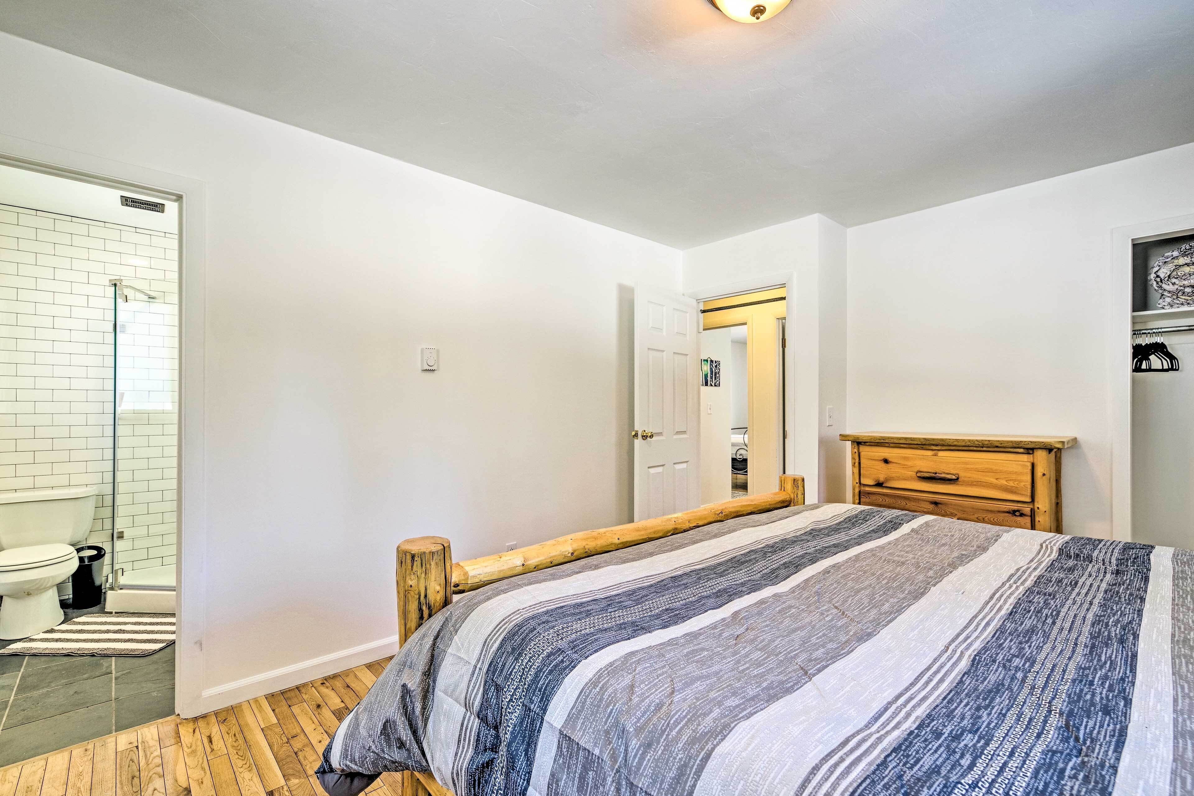 A bedroom with a wooden bed frame, a striped bedspread, a wooden dresser, and a white door leading to a bathroom with a white toilet and a glass shower enclosure.