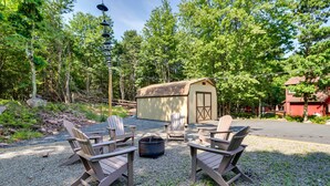 Et udendørs opholdsområde med Adirondack-stole og et bålsted, omgivet af træer og en lille skur.