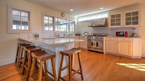A modern kitchen with a central island, wooden bar stools, white cabinetry, and a stainless steel range hood. - Luxe Waterfront East Quogue Home w/ Beach On-site! (East Quogue)