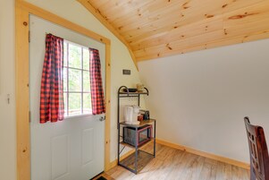 Una habitación con una puerta blanca, cortinas a cuadros rojos y negros, techo de madera, un estante metálico pequeño con un microondas, y un letrero que dice “MAINE”.