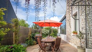 Une terrasse avec une table ronde en bois et des chaises, un parasol rouge et un mur en pierre.