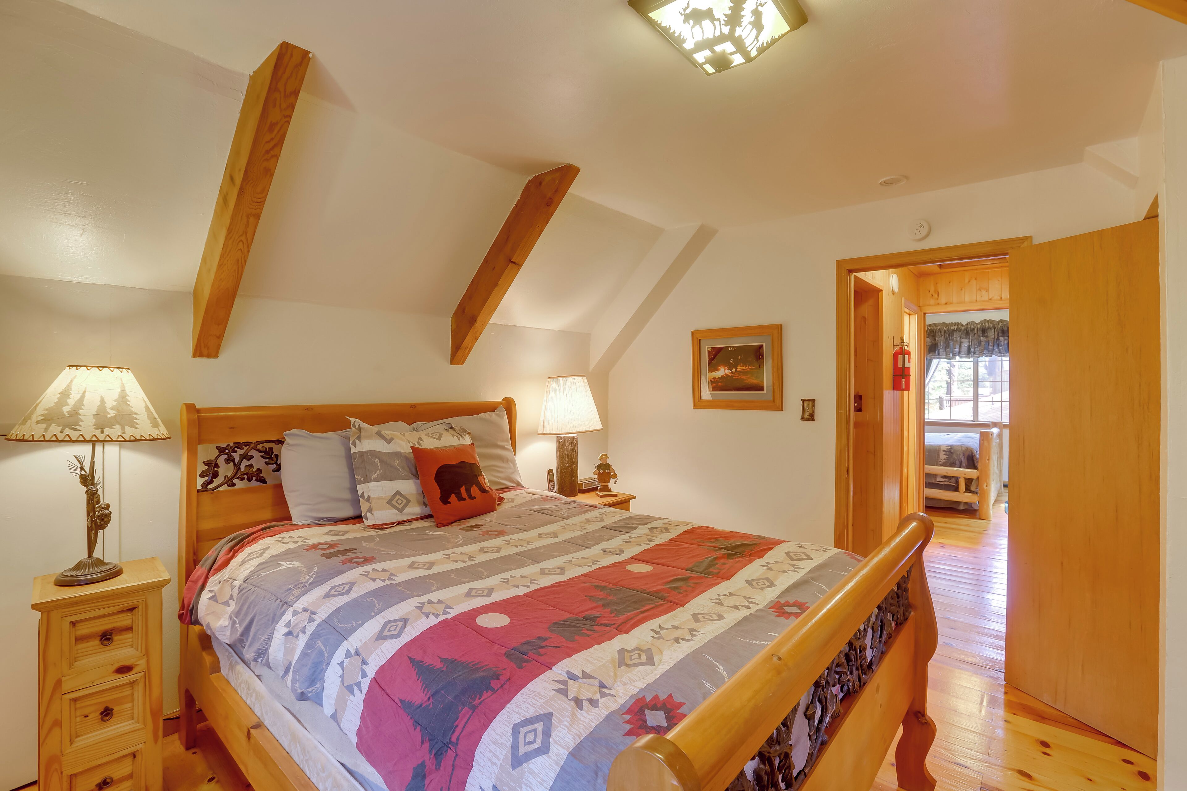 Un dormitorio con cama de madera, dos mesitas de noche con lámparas, techo con vigas de madera y una puerta que da a otra habitación.