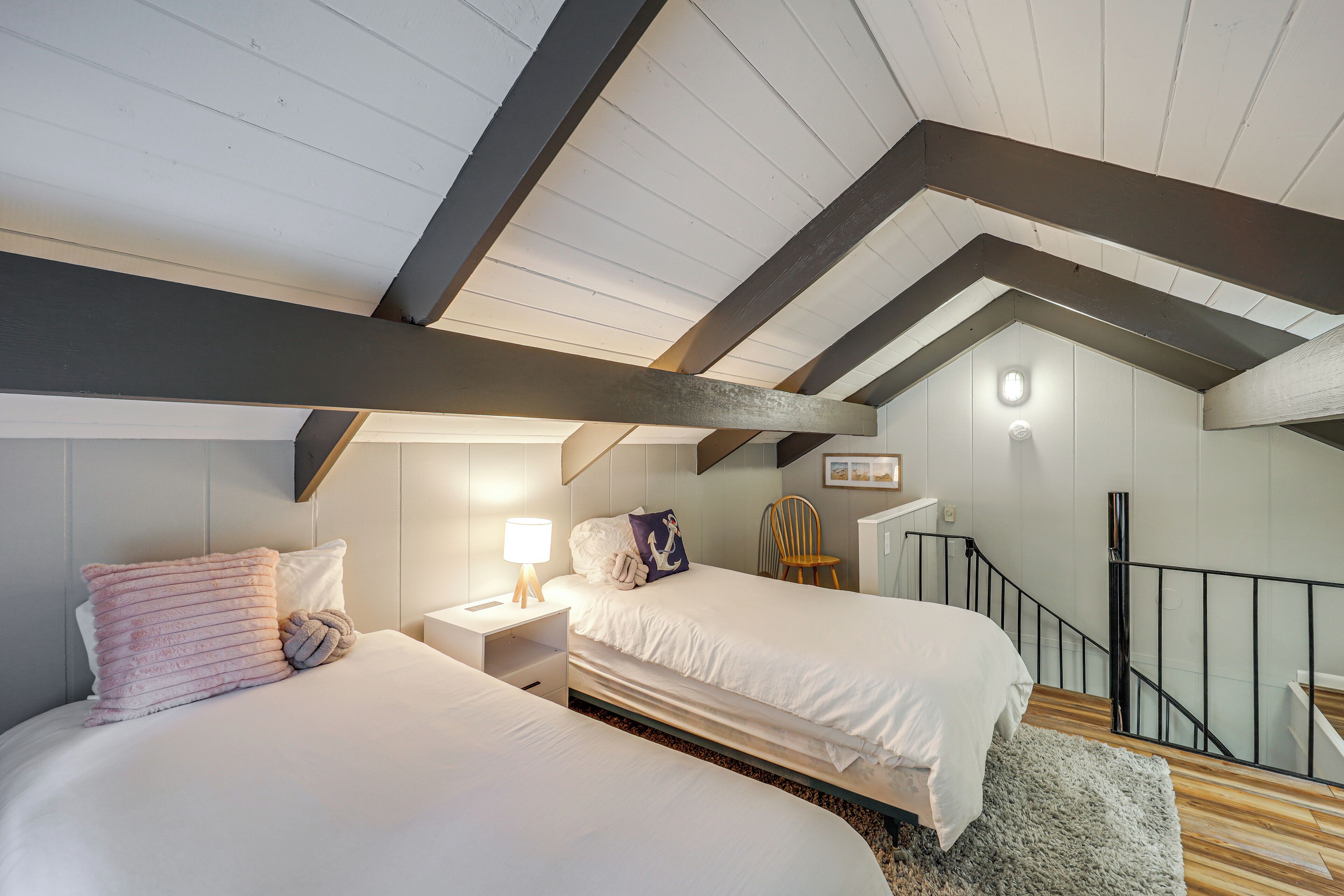 Quarto com duas camas, mesa de cabeceira com luminária, piso de madeira e escada com corrimão.