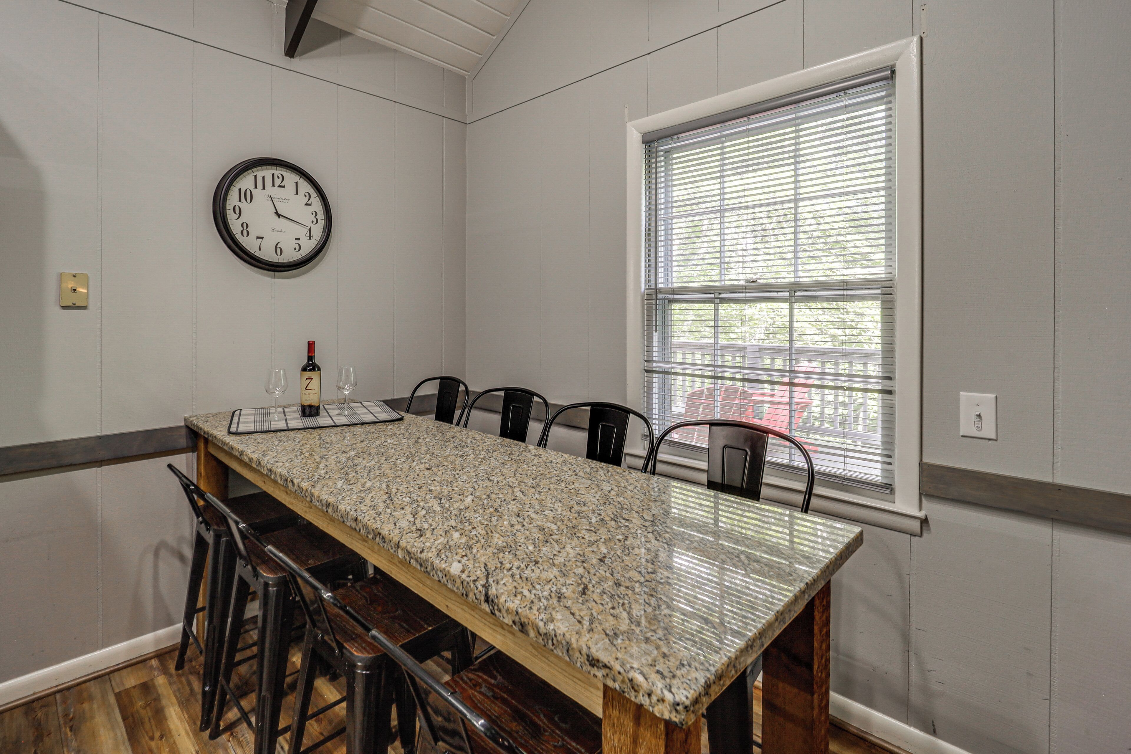 A dining room with a long table, chairs, a clock, and a window with blinds.
