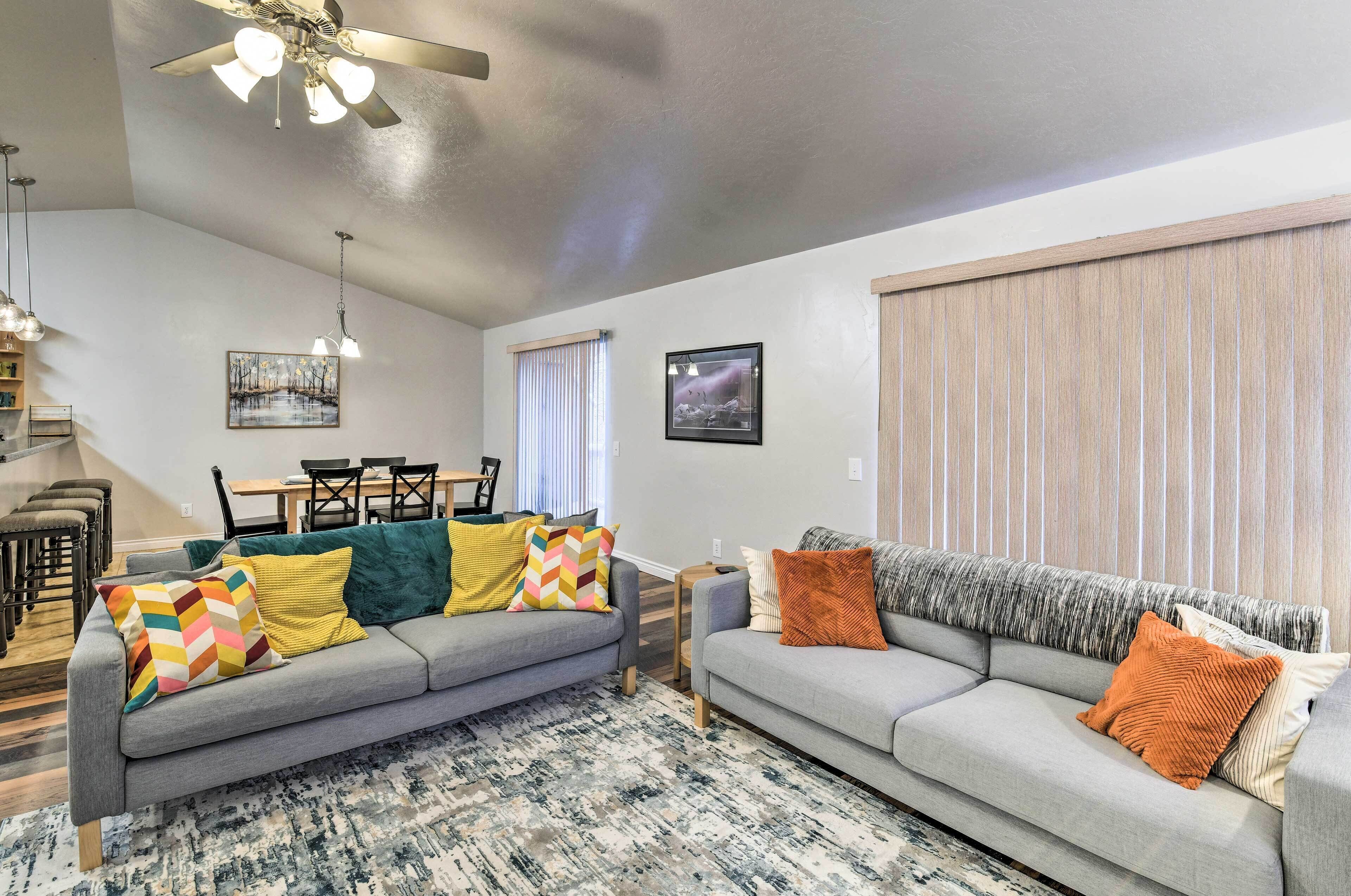 A living room with two grey sofas, colorful cushions, a ceiling fan, and a large window with blinds.