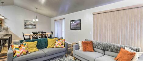 A living room with two grey sofas, colorful cushions, a ceiling fan, and a large window with blinds.
