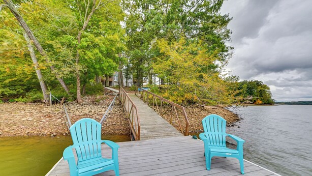 Une jetée en bois avec deux chaises Adirondack bleues, donnant sur une étendue d’eau, entourée d’arbres et reliée à un bâtiment par une passerelle en bois.