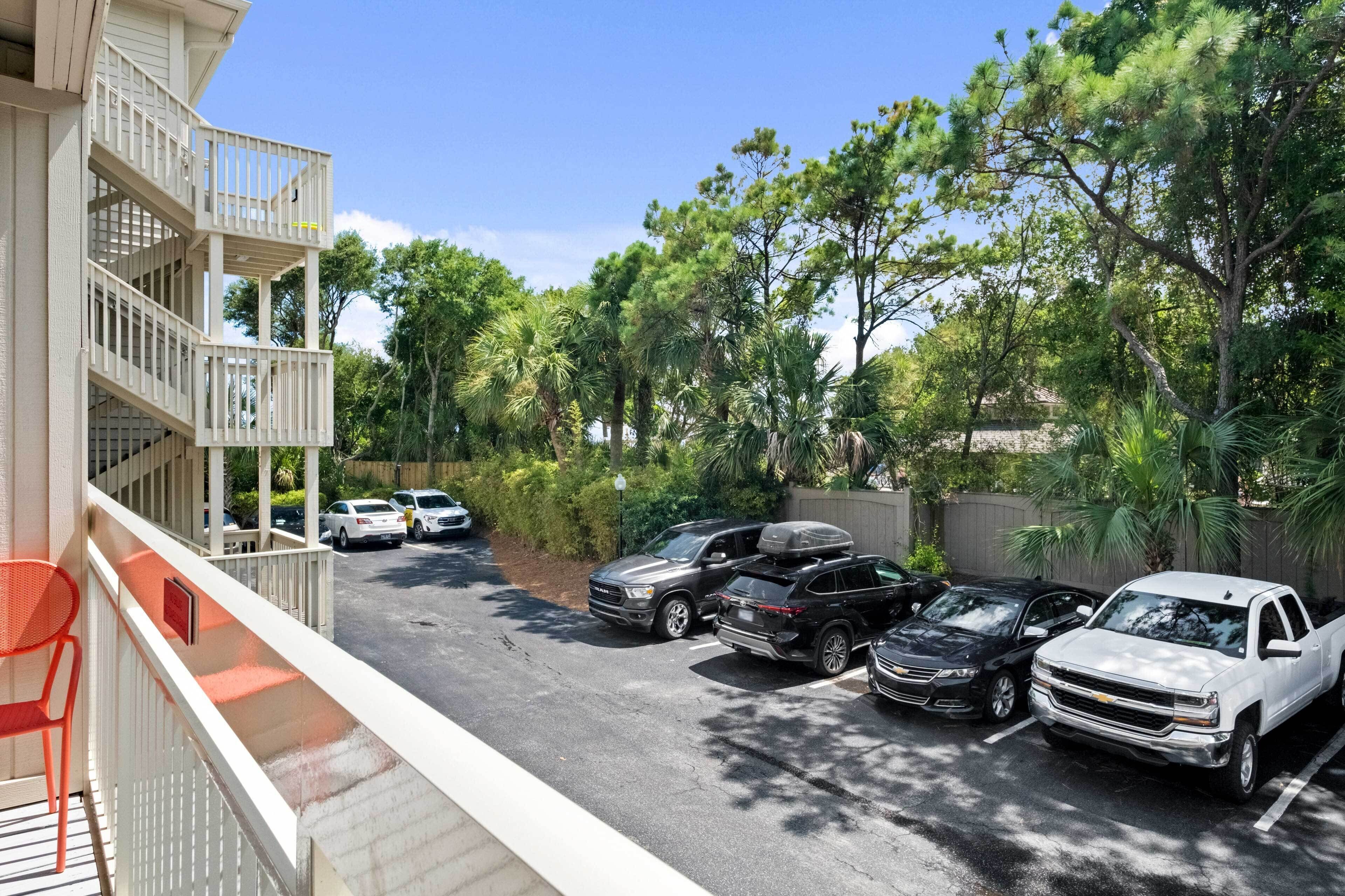 A parking area with several cars, a balcony with a chair, and trees in the background.