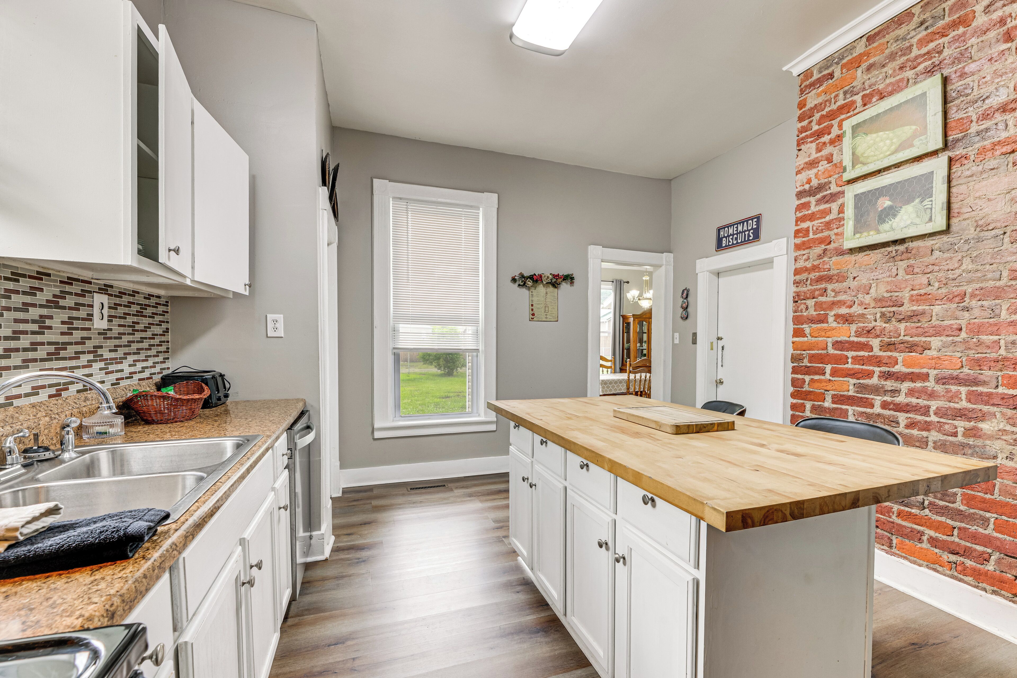 A kitchen with a granite countertop, white cabinets, a brick accent wall, and a view of the outdoors.