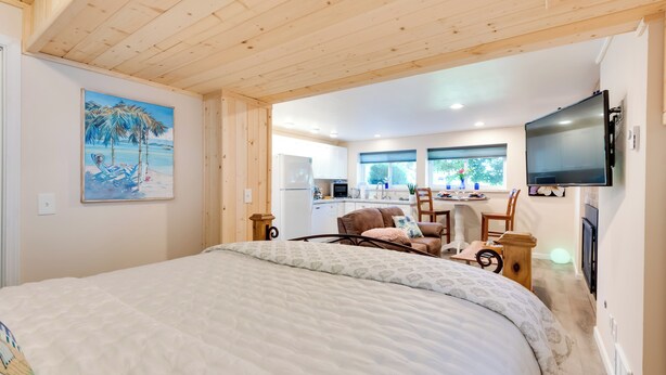 Une chambre avec un grand lit, un tableau représentant une scène de plage, une kitchenette et un téléviseur fixé au mur.