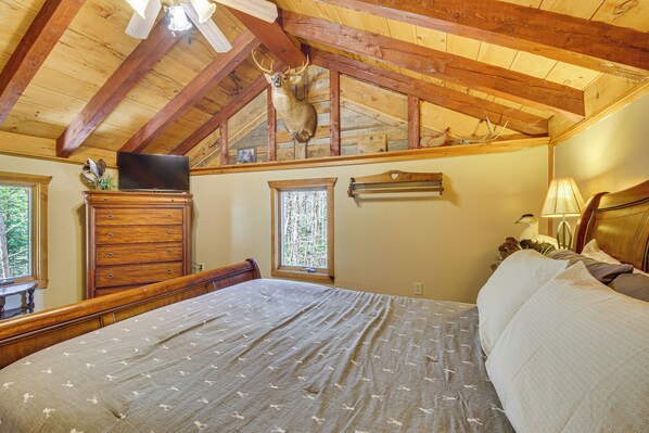 Un dormitorio con una cama grande, vigas de madera en el techo, una cabeza de ciervo montada en la pared, un aparador, un televisor y una ventana.
