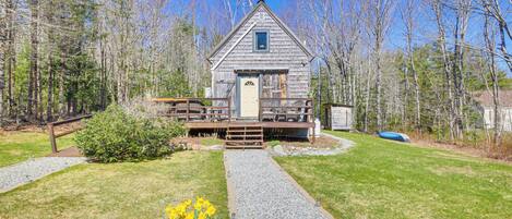 A rustic wooden cabin with a gravel pathway leading to the entrance, surrounded by a forest.