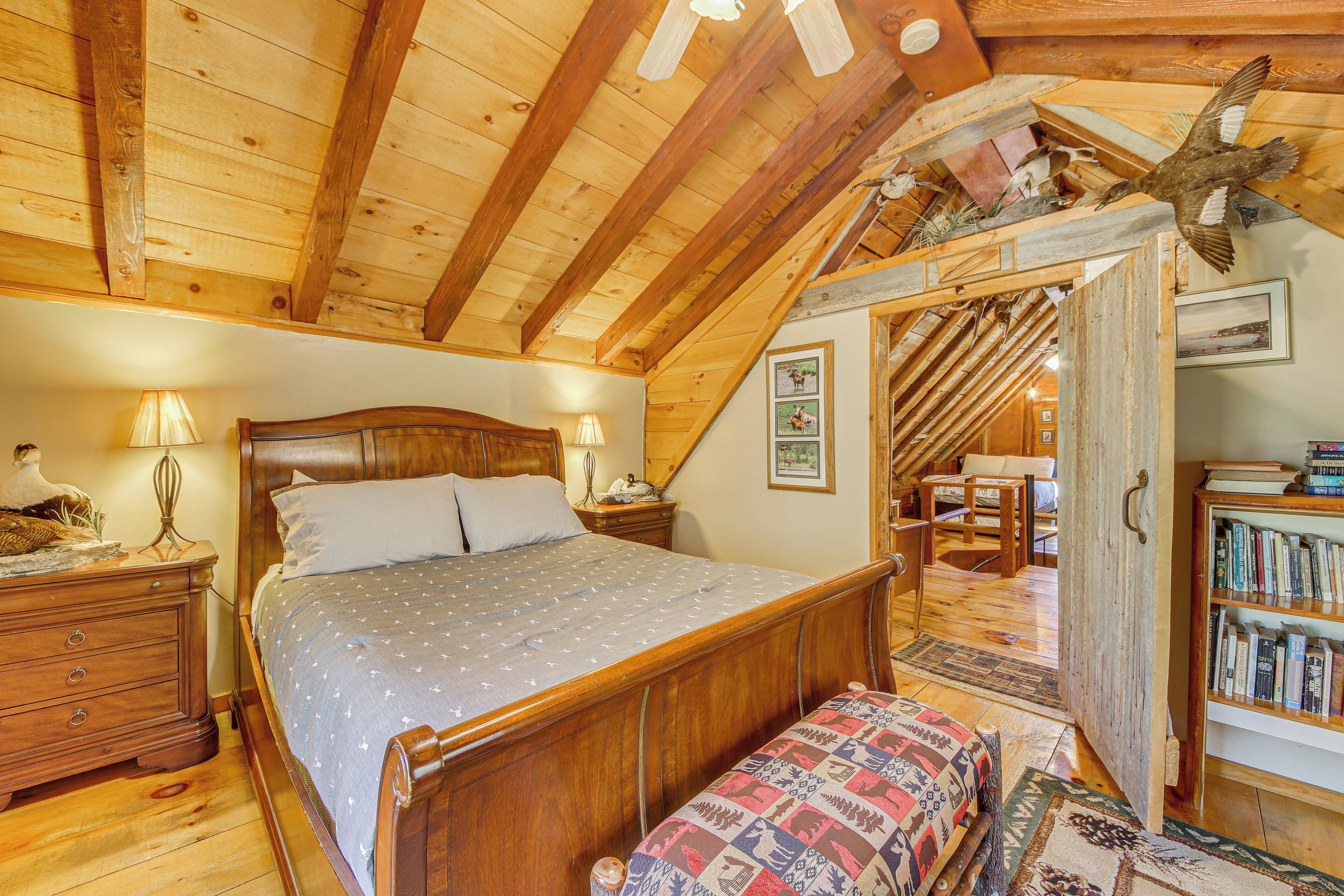 Un dormitorio con techo de madera, una cama con colcha estampada, una mesita de noche, una lámpara, una estantería y una puerta que da a otra habitación.