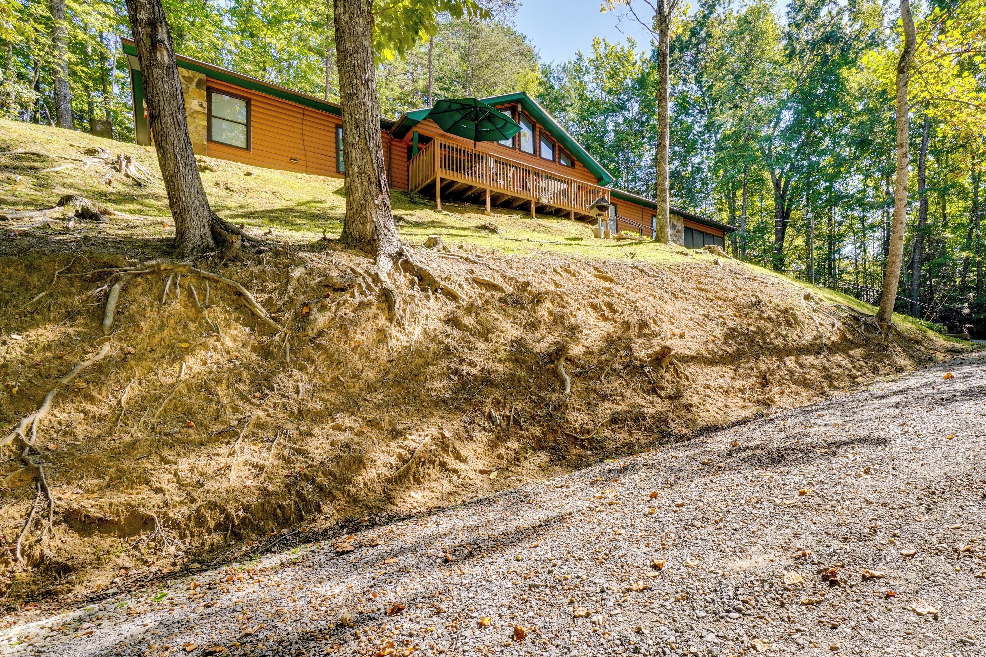 A cabin with a wooden deck and a green roof, surrounded by trees and a sloped landscape.
