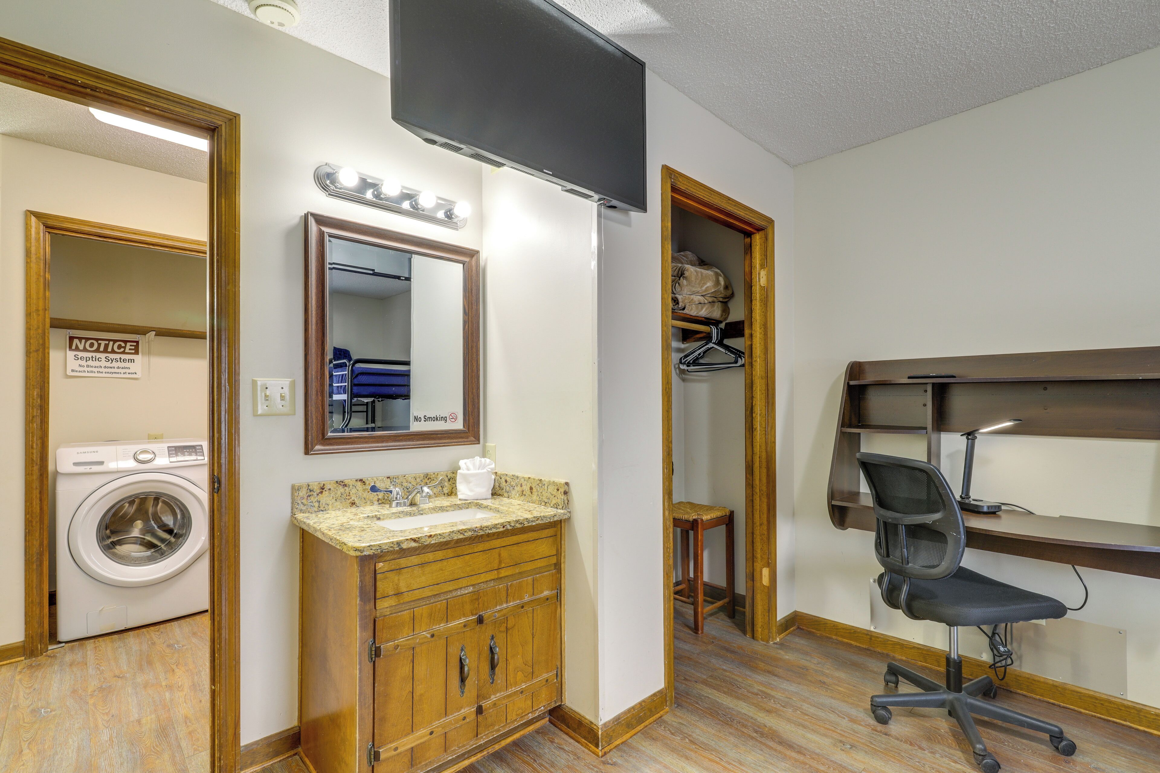 A compact hotel room with a washing machine, a sink with a vanity, a desk with a chair, and a television mounted on the wall.