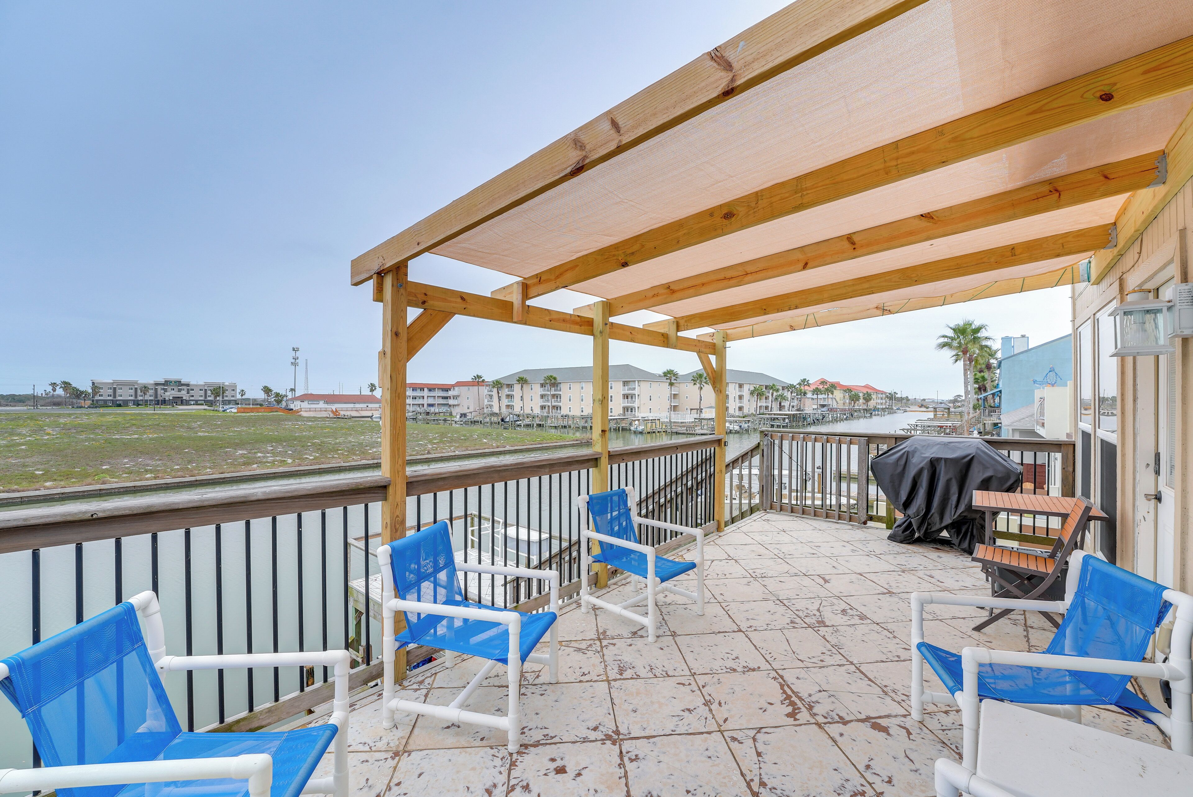 A covered outdoor seating area with blue chairs and a grill, overlooking a waterfront with buildings in the background.