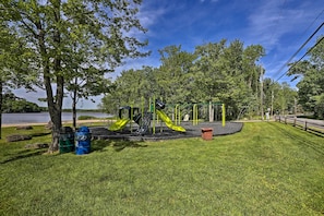 A playground with slides and climbing structures, surrounded by grass and trees, with a body of water in the background.