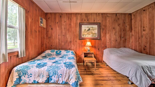 A room with wooden walls, a bed with a floral bedspread, a bedside table with a lamp, and a framed picture on the wall. - Lake Retreat w/ Dock, Deck & Bbq, BYO Boat! (Dent)