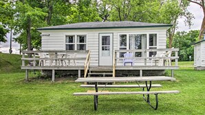 A small white cabin with a porch, a picnic table, and several Adirondack chairs. - Lake Retreat w/ Dock, Deck & Bbq, BYO Boat! (Dent)
