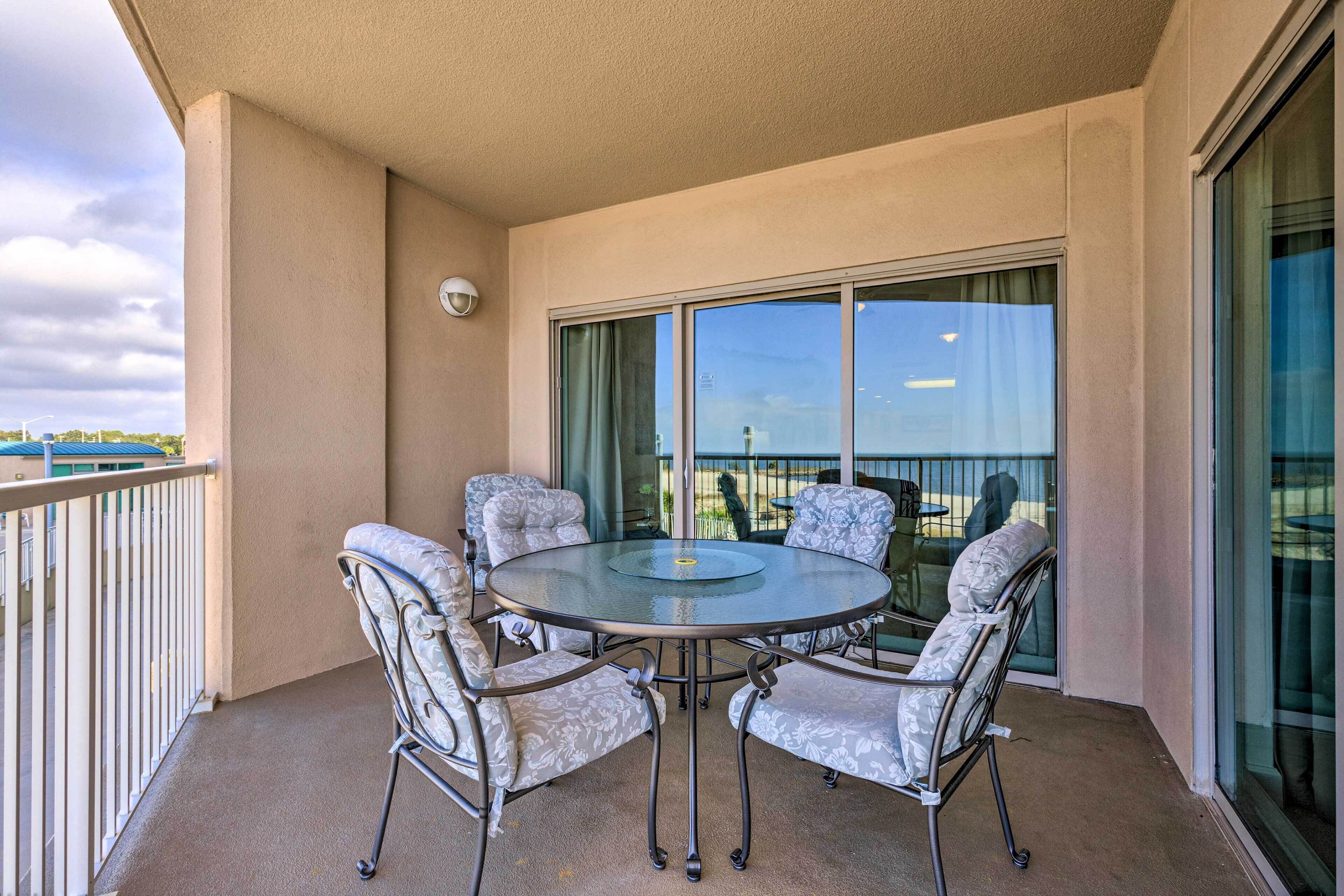 A balcony with a glass table and four chairs, large windows, and a view of a building and trees.