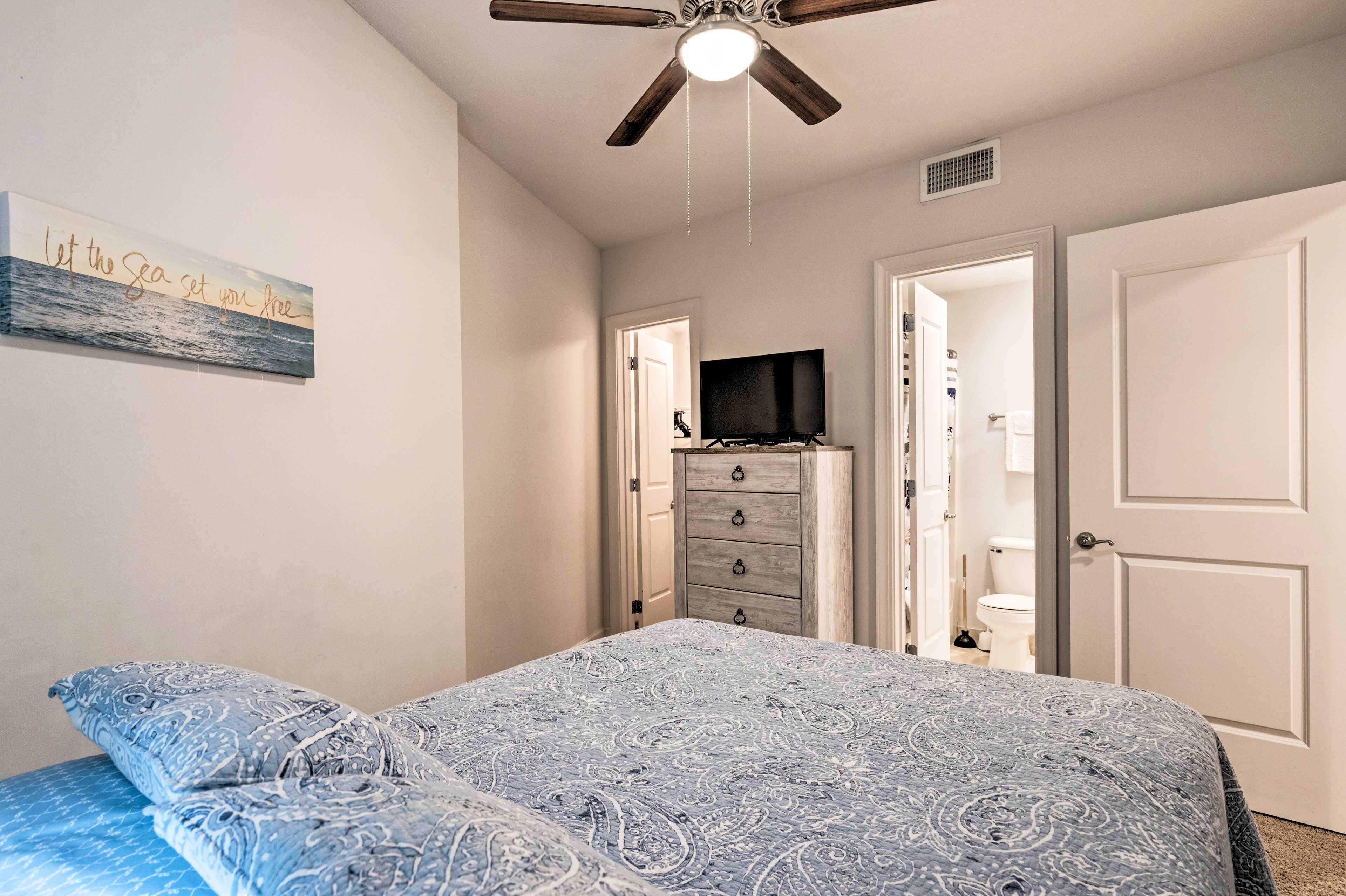 A bedroom with a bed, a ceiling fan, a dresser, a television, and a door leading to a bathroom.