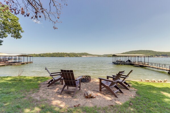 A lakeside setting with wooden chairs and a fire pit, surrounded by a dock and a grassy area.