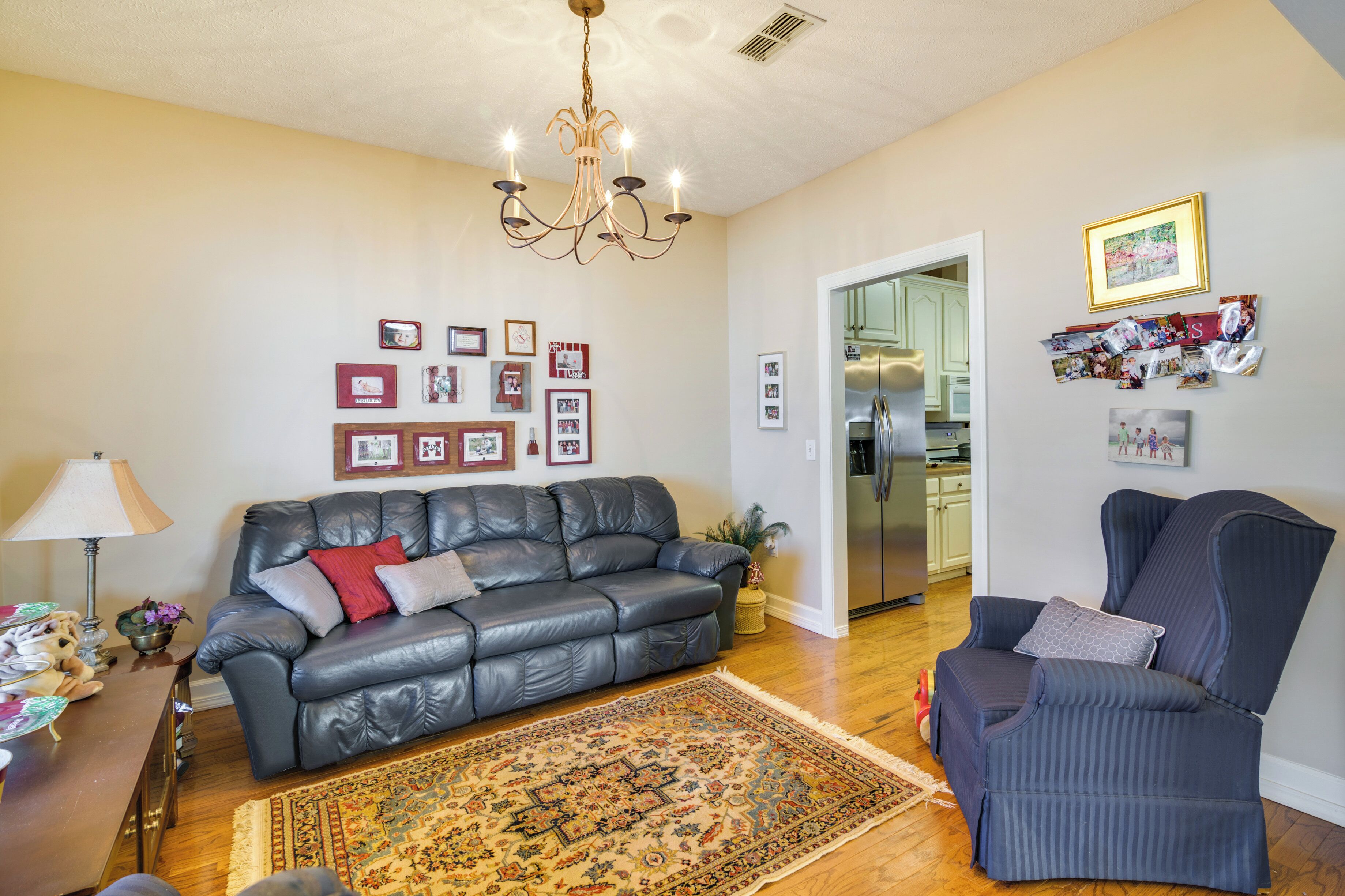 A living room with a black leather sofa, a patterned rug, a chandelier, and a wall adorned with framed photos.