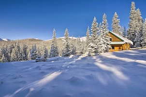 A log cabin with a snow-covered roof, surrounded by evergreen trees and a clear blue sky.
