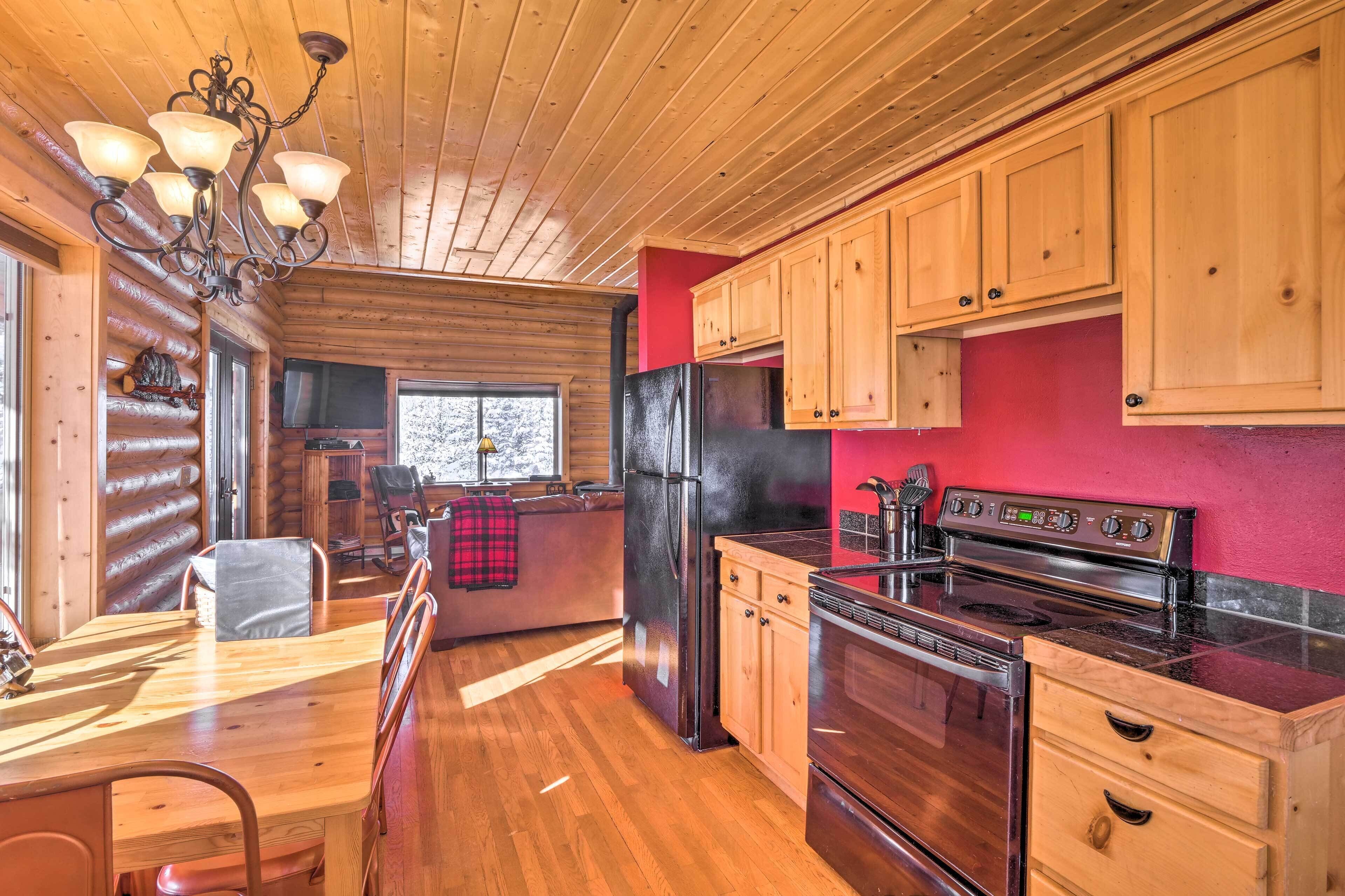 A log cabin interior with a wooden dining table, chairs, a kitchen with wooden cabinets, a refrigerator, a stove, and a chandelier.