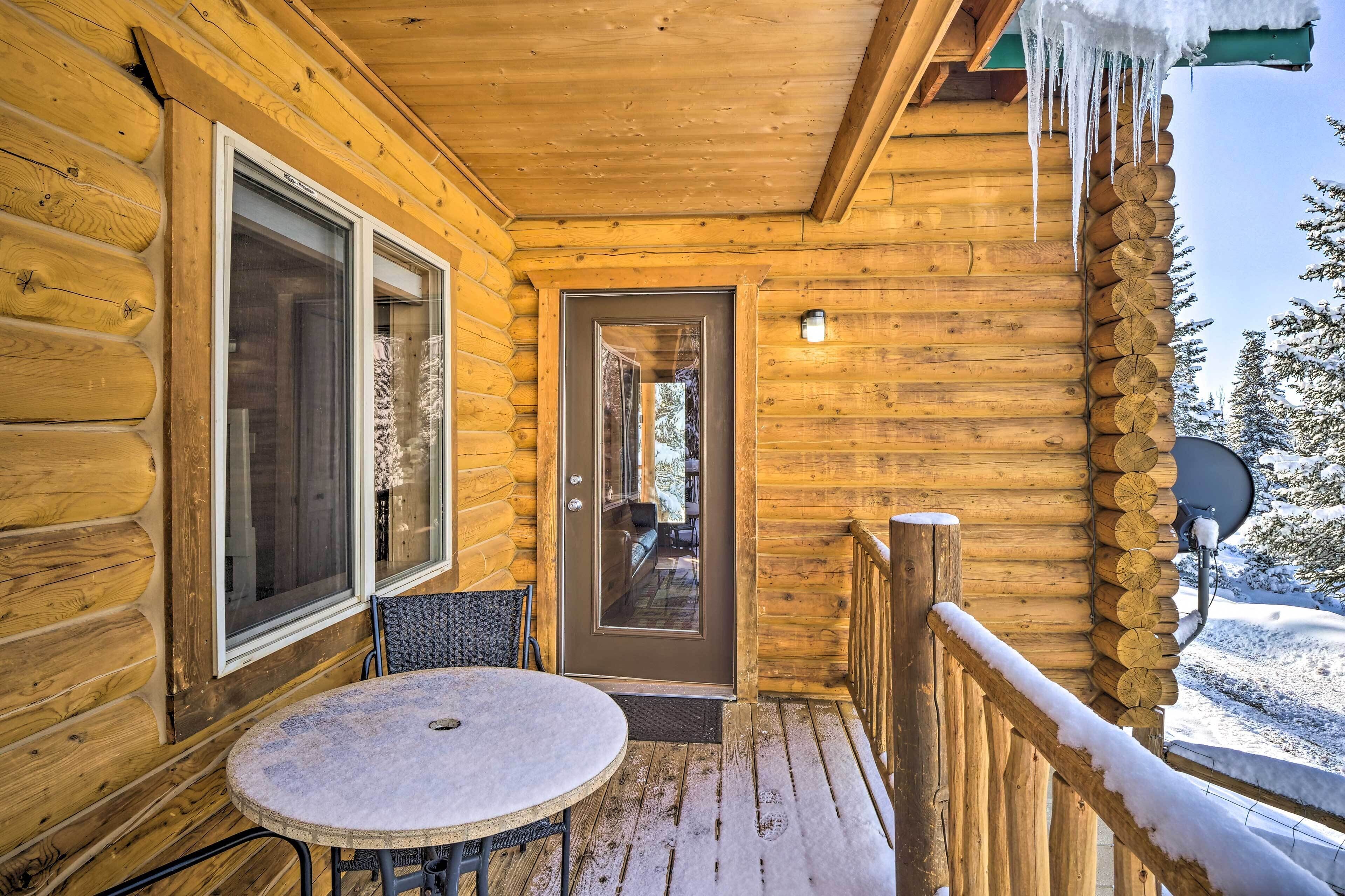 Una cabaña de madera con un porche cubierto de nieve, una puerta que da al interior, y una mesa redonda con sillas.