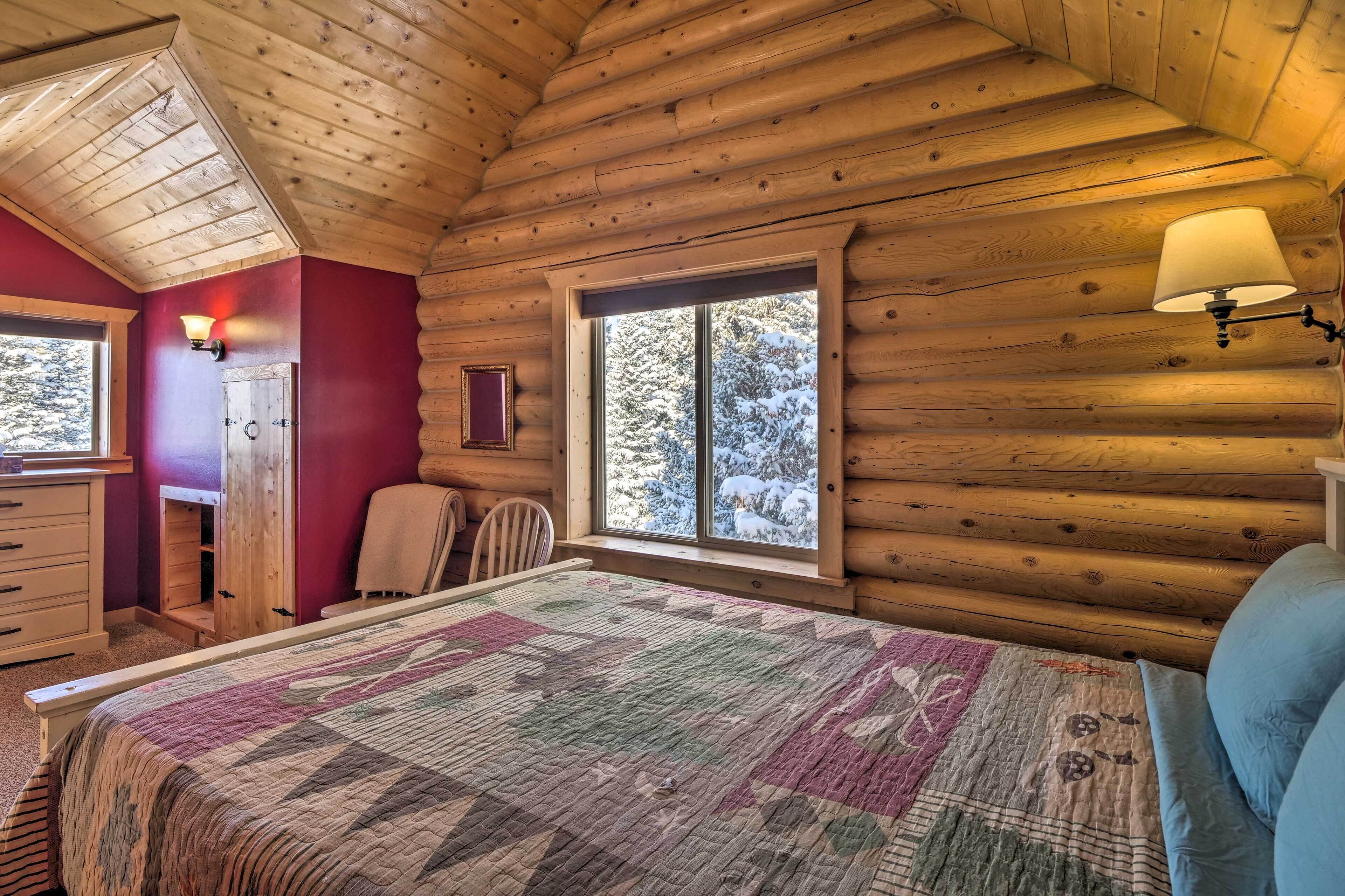 Une chambre à coucher dans un chalet en rondins, avec un lit, une commode, une chaise et une fenêtre donnant sur des arbres couverts de neige.
