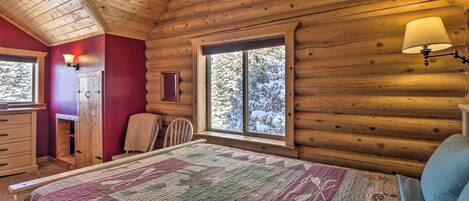 Une chambre à coucher dans un chalet en rondins, avec un lit, une commode, une chaise et une fenêtre donnant sur des arbres couverts de neige.