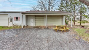 A single-story building with a garage door, a small porch with a wooden table, and a gravel driveway. - Central & Private Milan Home ~ 10 Mi to Sandusky! (Milan)