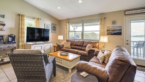 A living room with a brown leather sofa, a wicker chair, a coffee table, a flat-screen TV, and a view of the outdoors through large windows. - Rutledge Hilltop Home on Cherokee Lake w/ Decks! (Rutledge)