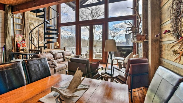 Un intérieur chaleureux avec une table à manger en bois, des fauteuils en cuir, un escalier en colimaçon et de grandes fenêtres offrant une vue sur l’extérieur.