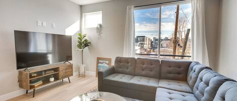 A modern living room with a large flat-screen TV, a grey sectional sofa, a glass coffee table, and a wooden TV stand.