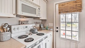 Una cocina con armarios blancos, un microondas, una estufa y un ventanal con persiana de madera.