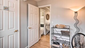 A hallway with a white door labeled "BATHROOM," a metal rack with baskets, and a wall-mounted lamp. - Bright Belmar Cottage < Half Mi to Boardwalk! (Belmar)