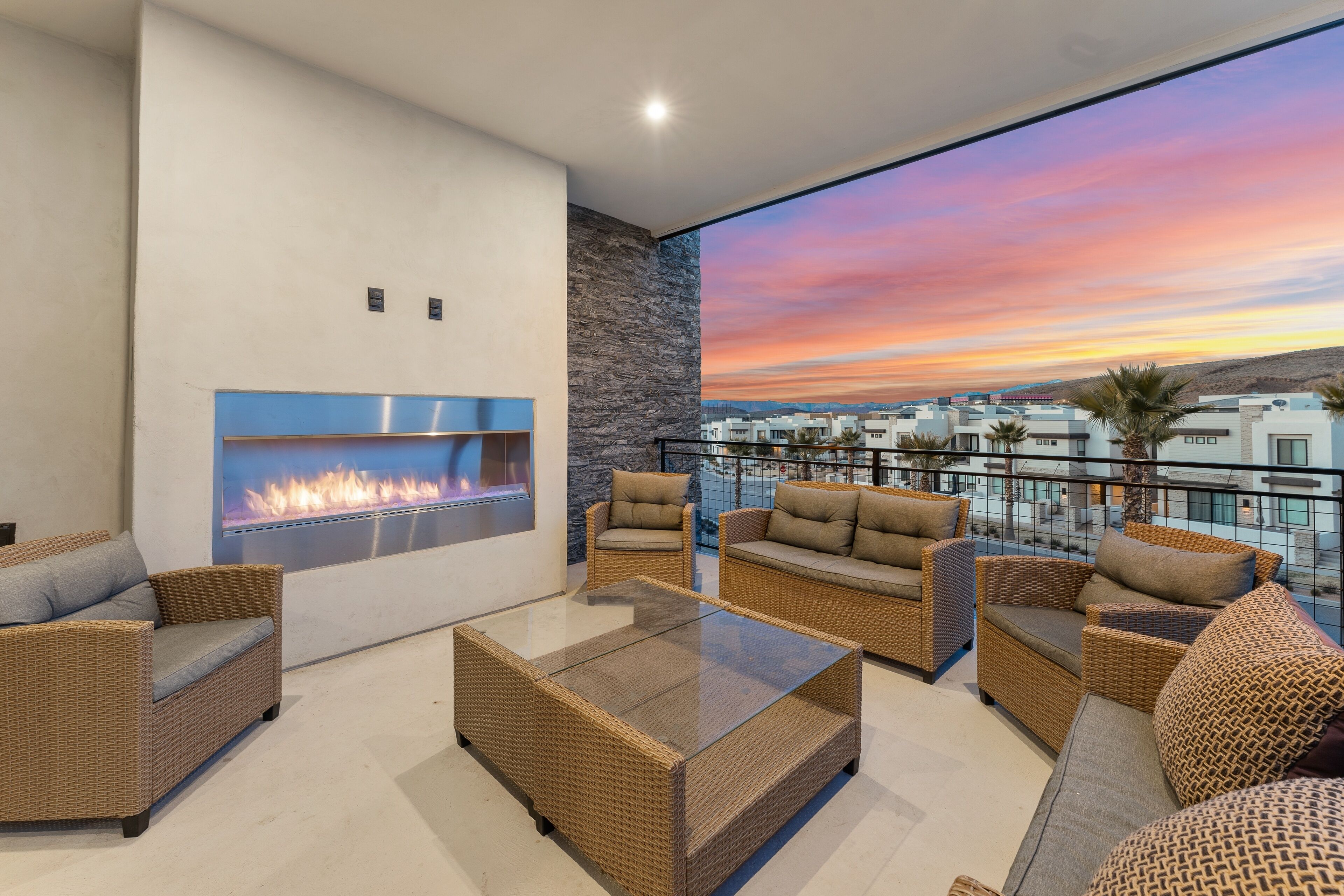 Un espacio exterior moderno con mobiliario de mimbre, una mesa de centro de vidrio y una chimenea integrada.