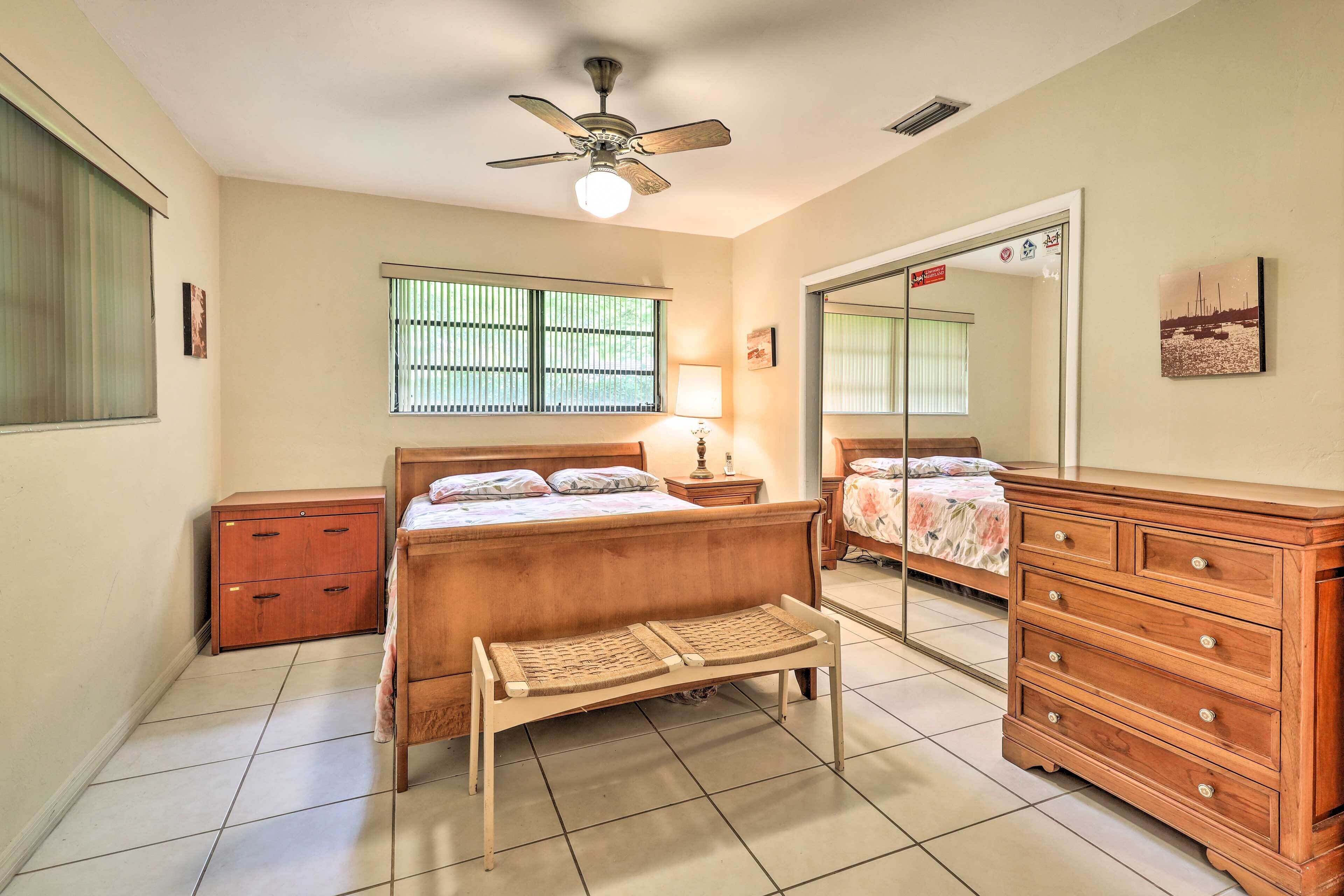A bedroom with a bed, a wooden bench, a dresser, a nightstand, a ceiling fan, and a window with blinds.