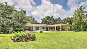 Une maison à deux étages, avec un toit recouvert de tuiles rouges, entourée d’une végétation luxuriante et de palmiers.
