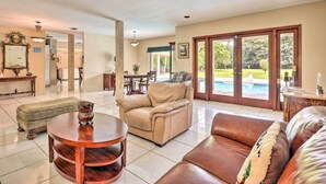 Un salon spacieux avec un canapé beige, un divan en cuir marron et une table basse ronde en bois. On y trouve également un coin repas avec une table et des chaises, ainsi qu’une grande fenêtre donnant sur une piscine.