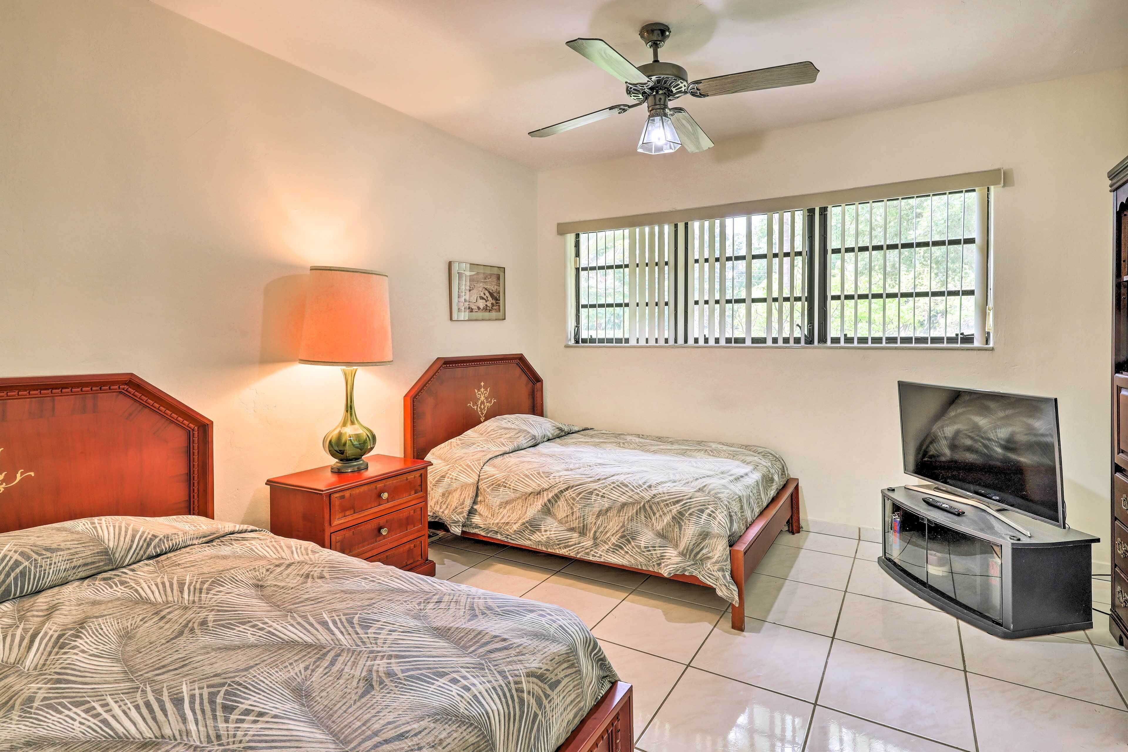 A bedroom with two beds, a TV stand, a ceiling fan, and a window with bars.