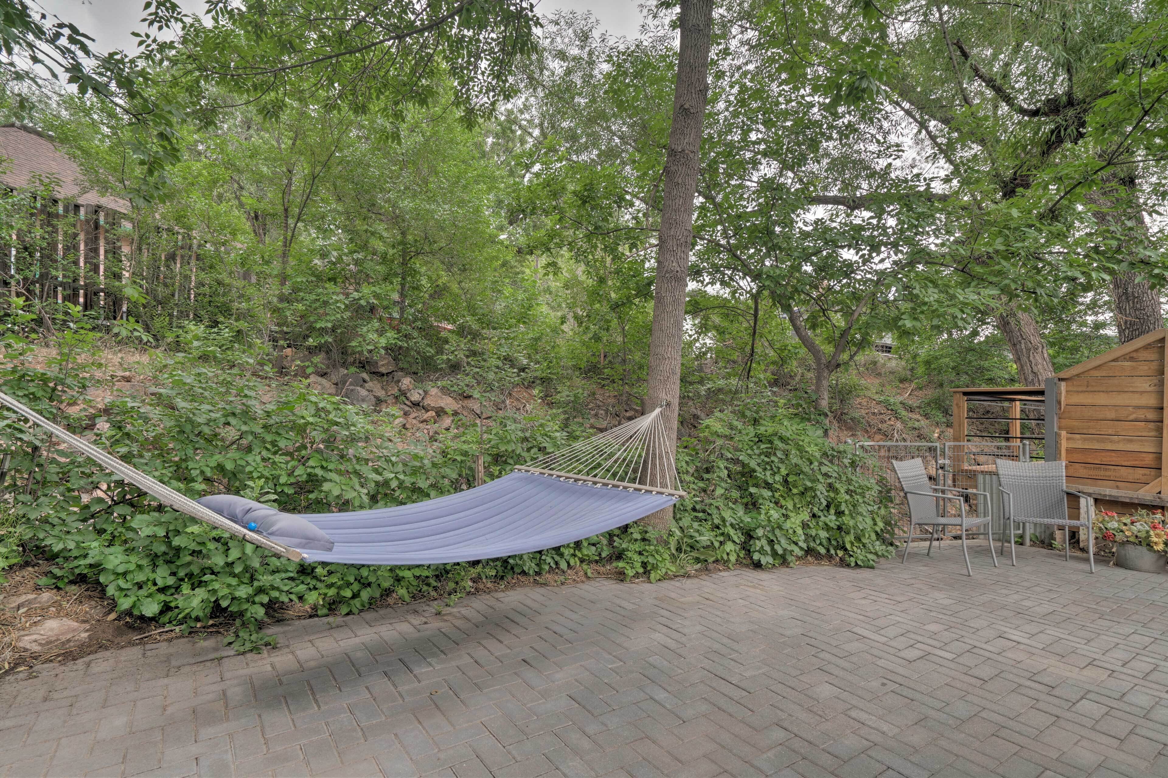 A hammock in a garden area with a brick pathway, surrounded by greenery and a wooden structure.