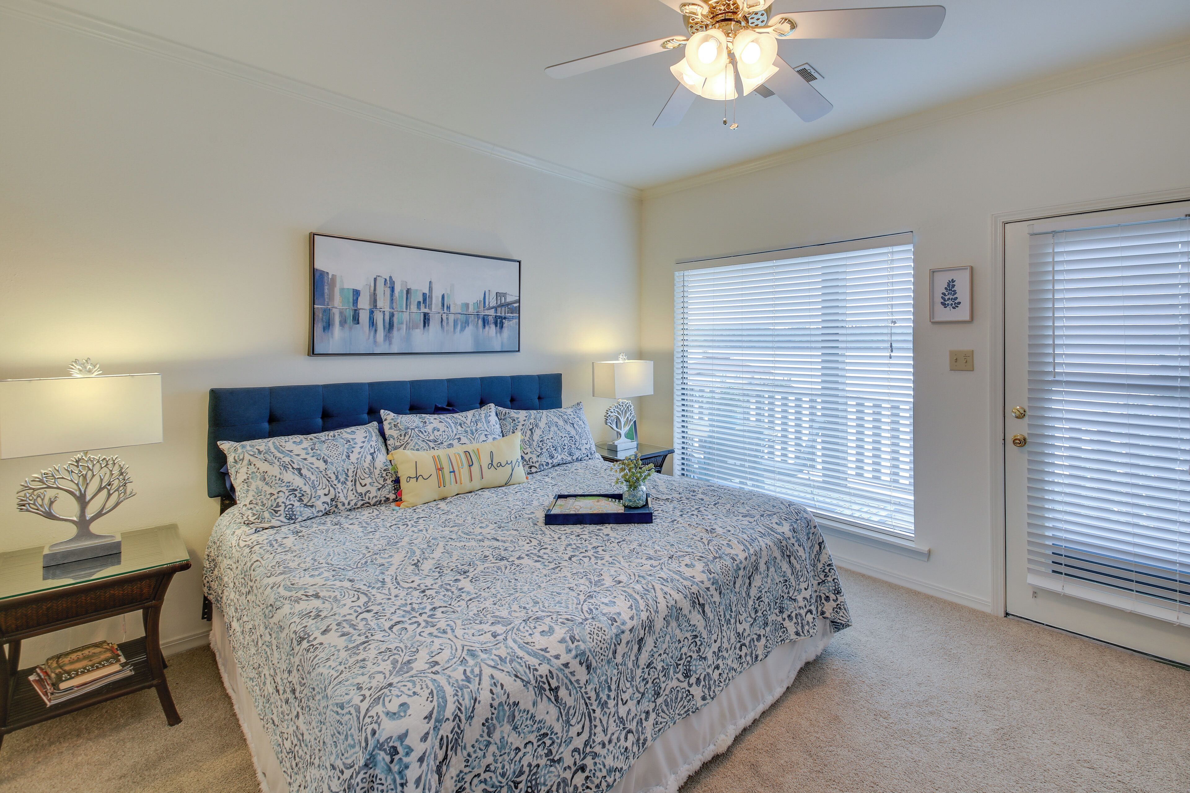 A bedroom with a large bed, a ceiling fan, a cityscape painting, and a door with blinds.